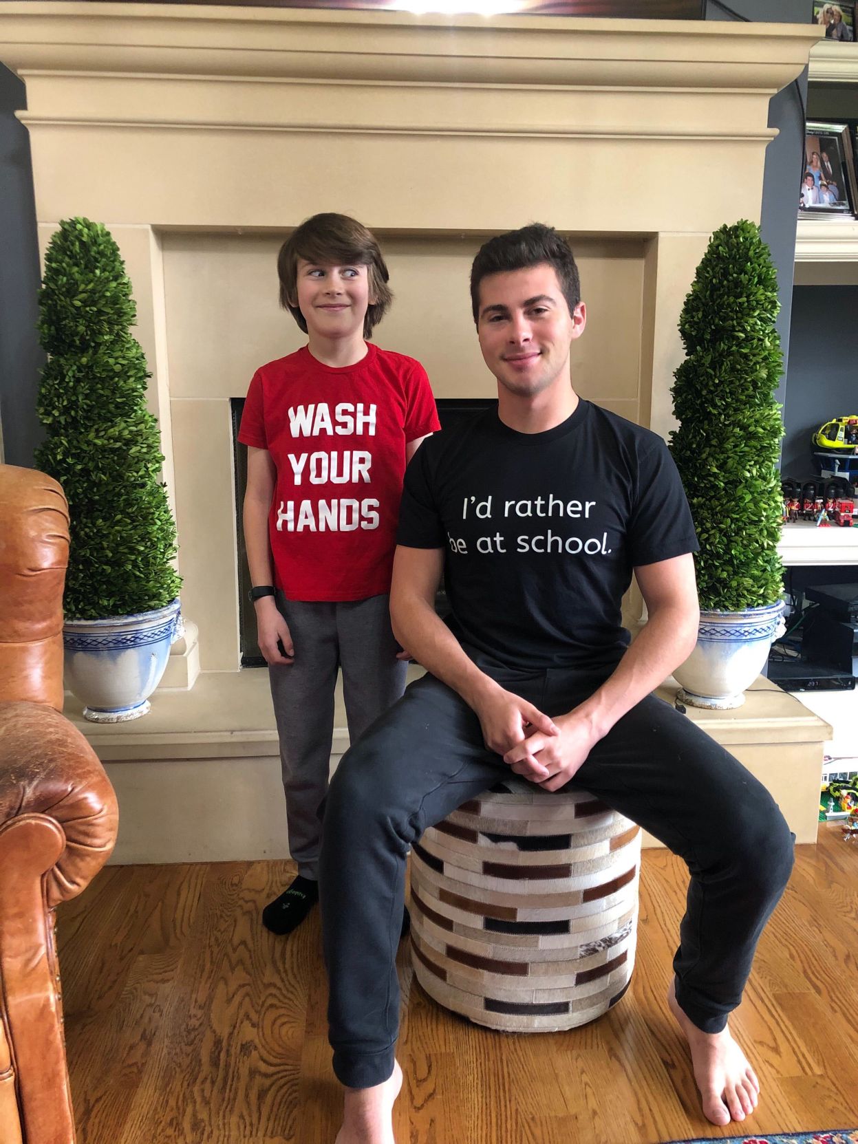 Two brothers smile at the camera. They wear t-shirts with the sayings, "Wash Your Hands" and "I'd rather be at school"